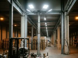 Des projecteurs LED industriels sur mât en acier éclairent une allée en béton d'un entrepôt logistique européen avec des rayonnages à palettes.