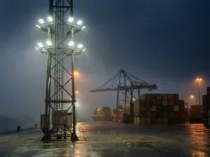 Mât d'éclairage industriel sur un quai en béton éclairant un terminal à conteneurs au crépuscule, avec une grue portique et des conteneurs empilés en arrière-plan.